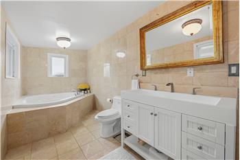 Bathroom featuring stone tile walls and flooring, a white dual-sink vanity with built-in cabinetry, and a jetted corner soaking