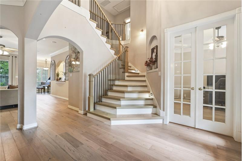 Open entryway with elegant wood flooring and a striking staircase. Arched doorways and French doors to office adds charm.