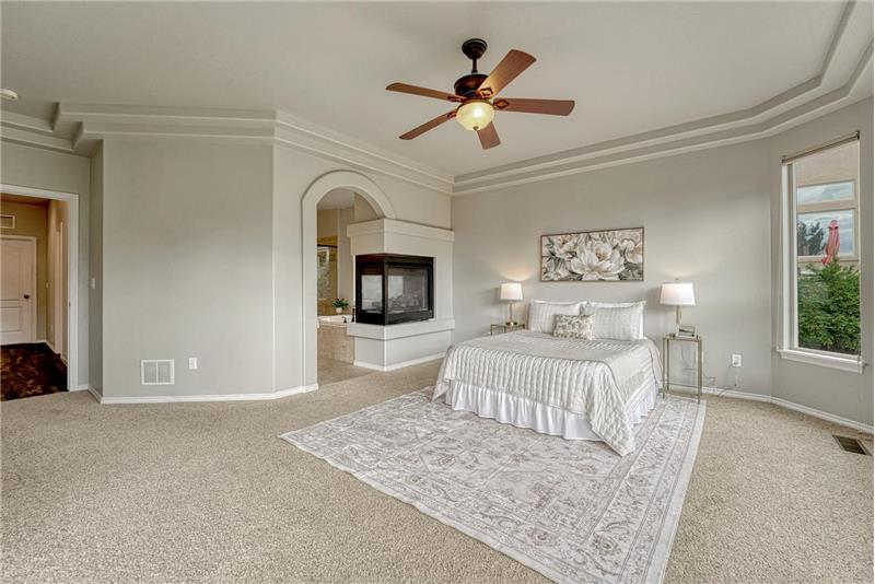 Relax in your private main level Primary Retreat with neutral carpet, a tray ceiling, lighted ceiling fan, and 3-side fireplace.